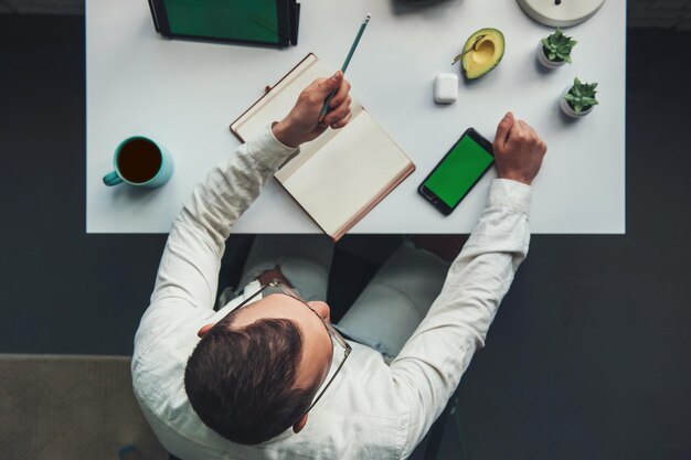 Businessman sitting in a chair at the office table writing in a copybook and using his phone top vie...