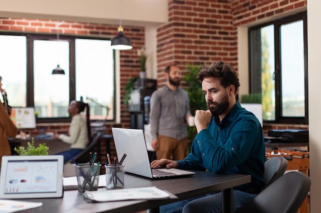 Businessman sitting at desk in startup office brainstorming ideas while working at marketing project analyzing company turnover using laptop. Diverse businesspeople planning business collaboration