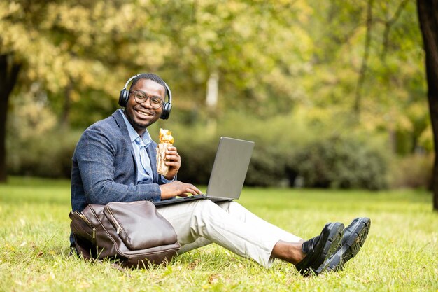 Businessman sitting on grass in the park eating his lunch and working on laptop