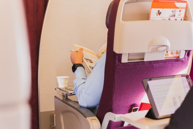 Businessman sitting on the plane reading magazine with cup of coffee.