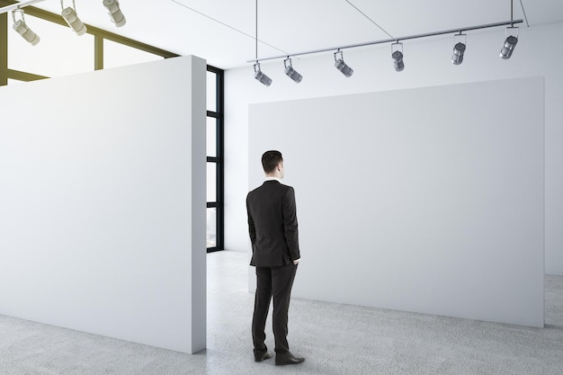 Businessman standing in clean gallery interior and looking on blank wall
