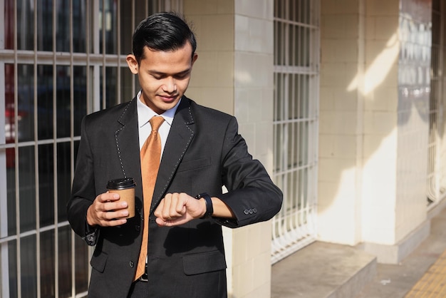 Businessman standing near office building with coffee cup and checking the time on watch