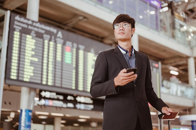 時間のフライトスケジュールの看板に立っているビジネスマンは、空港ターミナルゲートでスマートフォンを保持します。