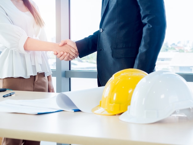 Businessman in suit, engineering or architect and woman shaking hands on blueprint and yellow and white safety hard hat on desk on glass window.