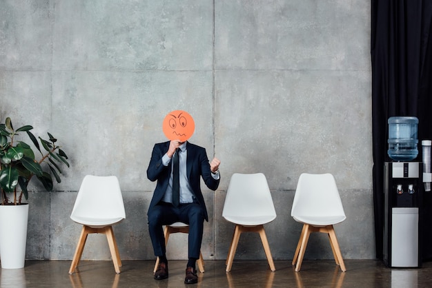 Businessman in suit sitting on chair and holding card with confused face expression in waiting hall