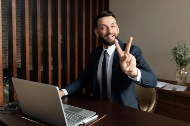 Businessman in a suit sitting in the office at a laptop showing a gesture of two fingers