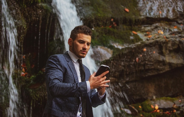 Businessman in suit with smartphone at waterfall background Waterproof device concept Telework business man with ip68 telephone