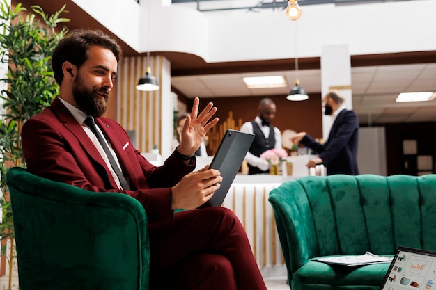 Businessman in suit working in lobby, attends videocall meeting while he is travelling abroad. Young adult arriving at hotel and using tablet to talk to colleagues, videoconference call.