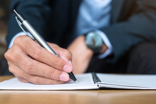 オフィスやカフェのノートに何かを書いているスーツのビジネスマン、紙のレポートに署名とペンを持っている男の手。ビジネスコンセプト