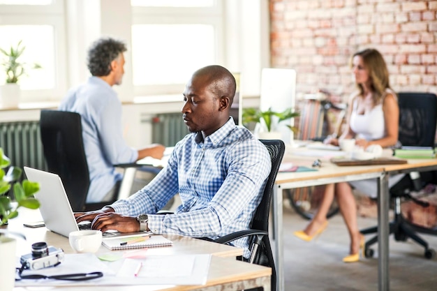 Businessman using laptop at desk in office with colleagues in background