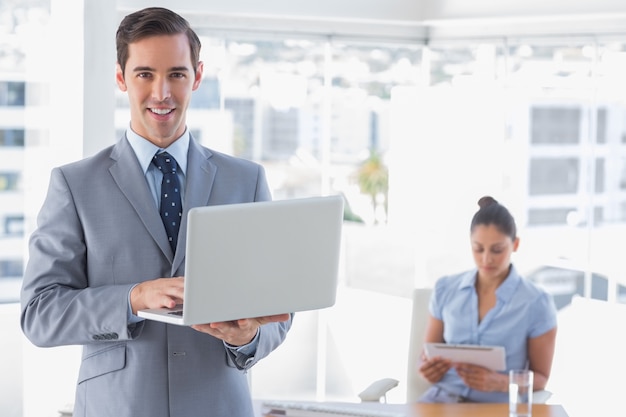 Businessman using laptop standing in office smiling at camera