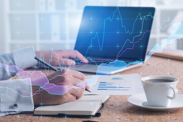 Businessman using laptop and writing in modern white office with double exposure of graph. Stock market concept. Toned image