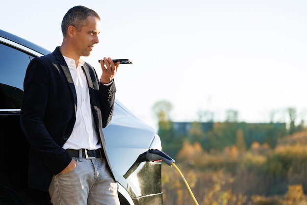 Businessman using smartphone voice messages and waiting power supply connect to electric vehicles