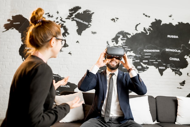 Businessman using virtual reality glasses sitting at the travel agency office with woman manager and world map on the background