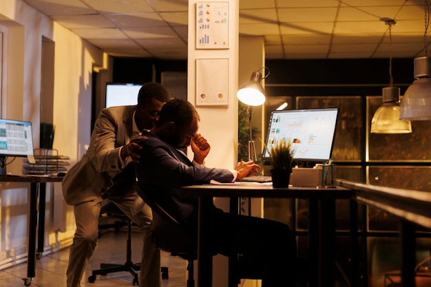 Businessman wakes up worker after falling asleep on table desk in startup office. African american entrepreneurs working overtime, planning financial investment strategy to increase company profit