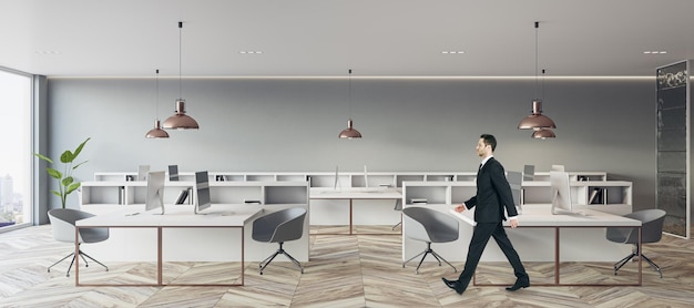 Businessman walking by white work table in spacious coworking\
office with wooden floor modern computers city view from panoramic\
window and light grey wall background