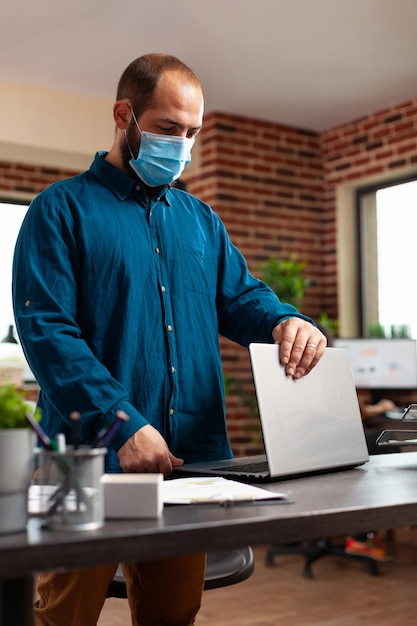 Photo businessman wearing medical protective face mask to prevent infection with coronavirus opening laptop working at company investment analyzing marketing strategy in startup office. business concept