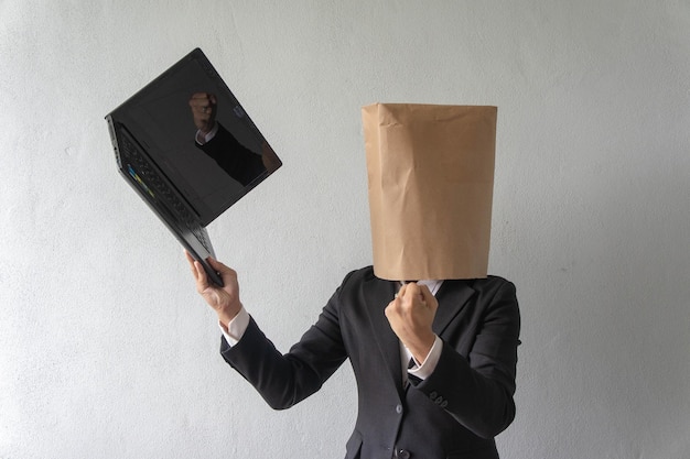 Photo businessman wearing paper bag over face while using laptop against wall