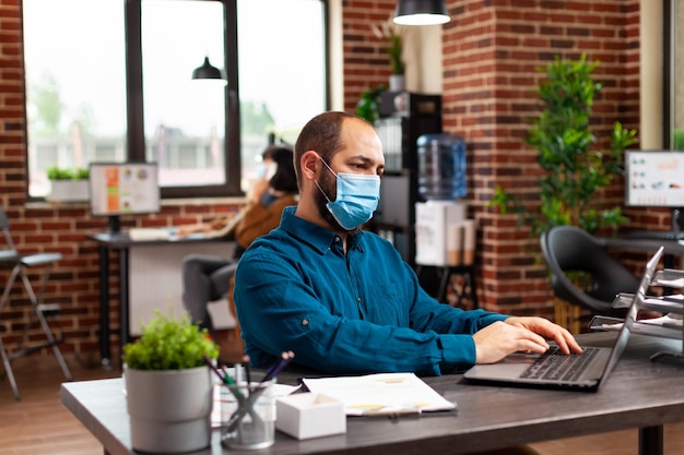 Uomo d'affari che indossa una maschera protettiva per proteggere l'infezione con la strategia di marketing di digitazione del coronavirus sulla tastiera del laptop che analizza il rapporto aziendale. manager che lavora alle statistiche finanziarie