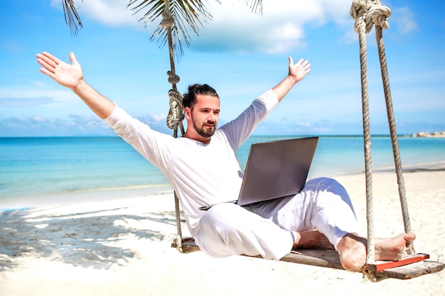 Бизнесмен, одетый в белый фрилансер, сидя на качелях на пляже с ноутбуком