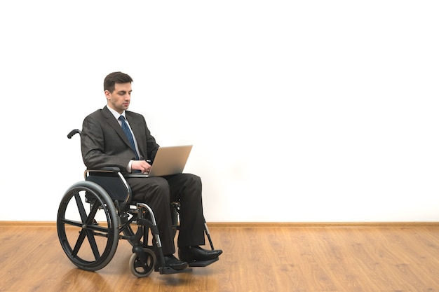 The businessman in a wheelchair sitting with a laptop on a white wall background