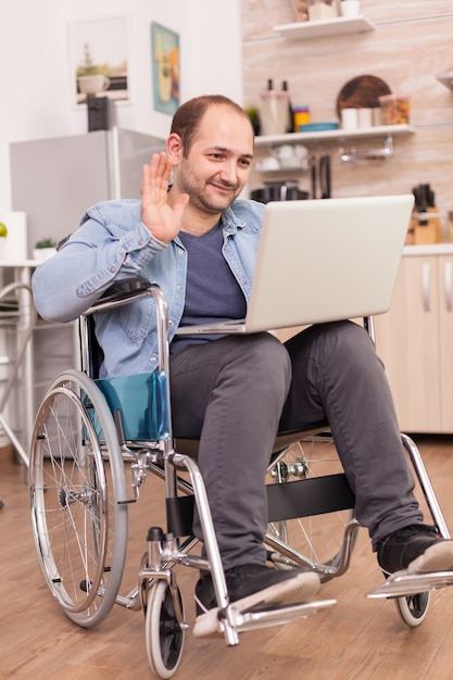 Uomo d'affari in sedia a rotelle che ondeggia durante una videoconferenza sul computer portatile in cucina mentre sta preparando il cibo. disabile paralizzato uomo handicappato con disabilità motoria che si integra dopo un incidente.