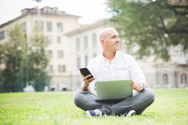 Uomo d'affari in camicia bianca che lavora con il computer portatile che si siede sull'erba in un parco