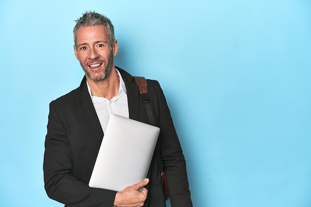 Businessman with backpack holding laptop on blue backdrop