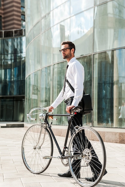 Businessman with bicycle. Full length of young businessman rolling his bicycle while walking outdoors