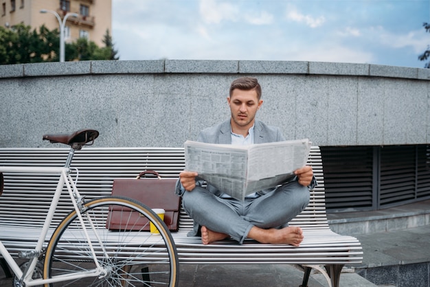 ダウンタウンのオフィスビルのベンチで新聞を読んで自転車を持つビジネスマン。