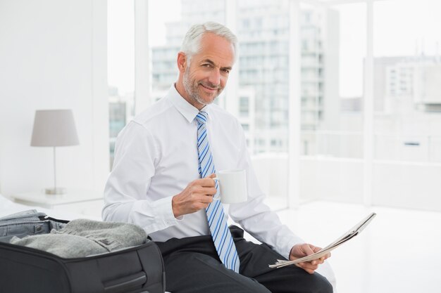 Businessman with coffee cup reading newspaper by luggage