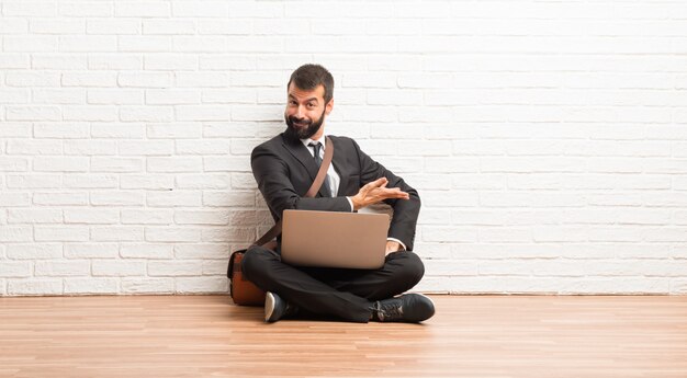 Businessman with his laptop sitting on the floor pointing back and presenting a product