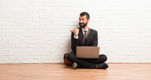 Businessman with his laptop sitting on the floor pointing to the side with a finger to present a product