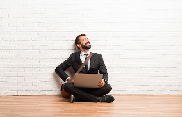 Uomo d'affari con il suo portatile seduto sul pavimento in posa con le braccia in anca e ridendo
