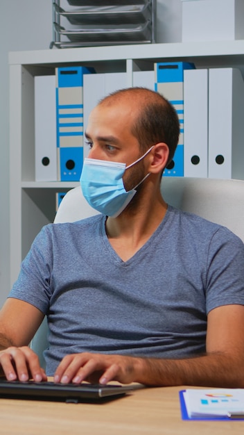 Businessman with medical protection face mask typing on computer in office room. Entrepreneur in new normal personal workplace corporate writing on pc keyboard looking at desktop