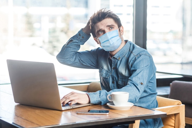파란색 셔츠를 입은 외과용 마스크를 쓴 사업가는 노트북에 앉아서 새로운 아이디어를 갖고 자신의 전략을 계획하고 한 손으로 머리를 잡고 있습니다. 실내 작업 및 건강 관리 개념입니다.