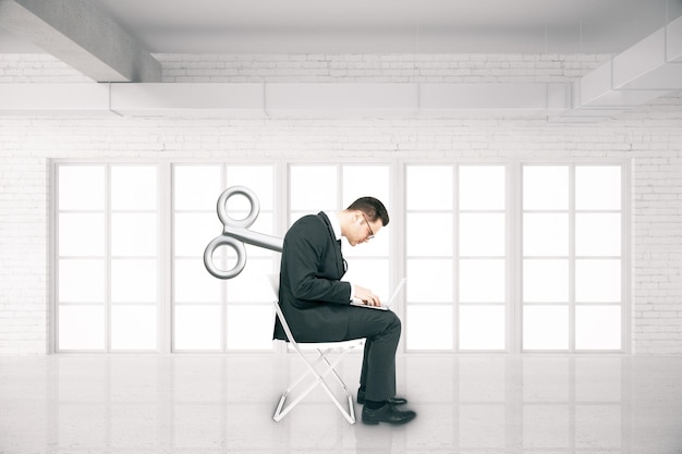 Бизнесмен с заводным ключом на спине сидит на стуле и пользуется ноутбуком в комнате из белого кирпича. Концепция контроля