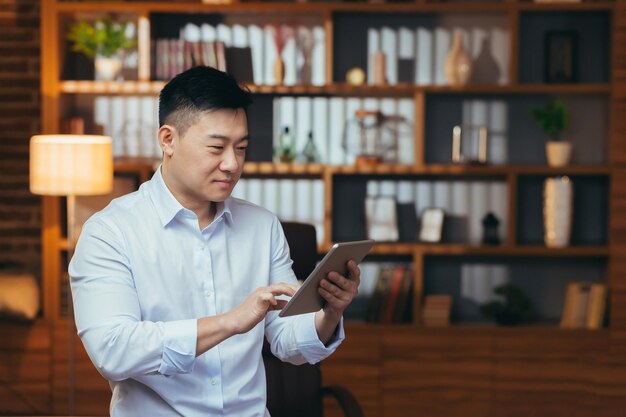 Businessman working on tablet in the evening man looks at the screen and smiles works late in the office happy asian corresponded on a dating site