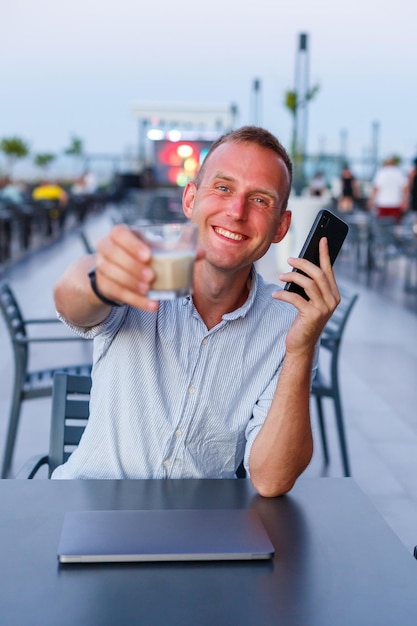 Uomo d'affari che lavora con il laptop sulla terrazza dell'hotel e parla al telefono per gestire i dipendenti durante le vacanze nel resort uomo in pantaloncini e maglietta che beve una bevanda rinfrescante