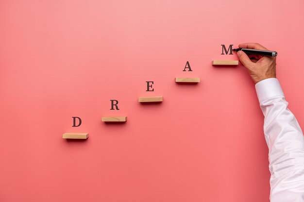 Photo businessman writing the word dream above wooden pegs placed in stairway like structure.