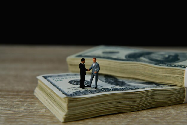 Businessmen figurine handshaking while standing on bundle of currency