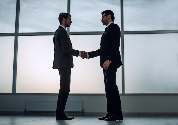 Businessmen shake hands standing near a large office window