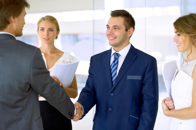 Businessmen shaking hands Two confident businessmen shaking hands and smiling while standing at office together with people in the background