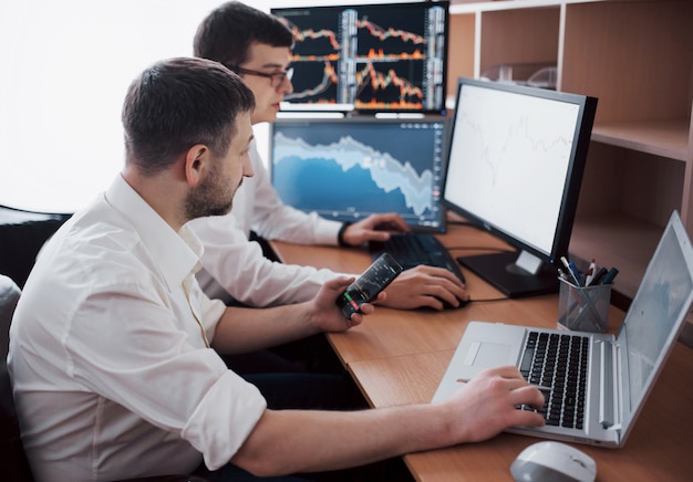 Businessmen trading stocks online. Stock brokers looking at graphs, indexes and numbers on multiple computer screens. Colleagues in discussion in traders office. Business success 