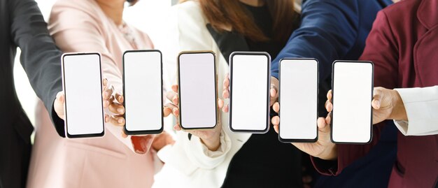 Businesspeople sitting together in office over working desk with colorful graph and charts pater, hold smartphones in different model with blank screens.