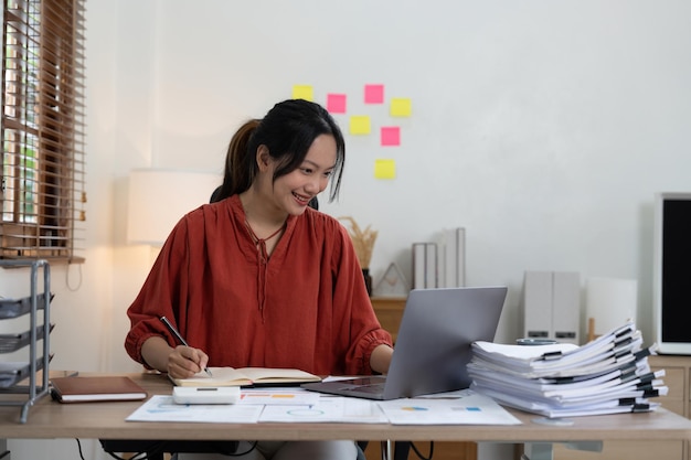 Businesswoman or accountant taking note and working on calculator and laptop computer to calculate business data during make note at notepad accountancy document at office