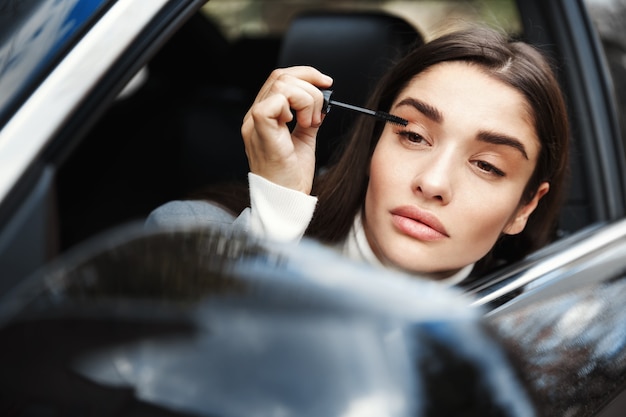 Businesswoman applying mascara and looking at rear-view mirror, put on makeup on her way to business meeting