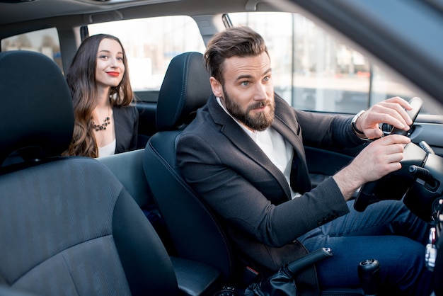 Imprenditrice sul sedile posteriore con un uomo elegante alla guida di un'auto in città. preoccupato per un ritardo in un incontro importante