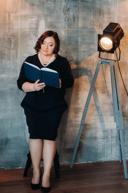 A businesswoman in a black beaded dress stands against the wall with a book in her hands