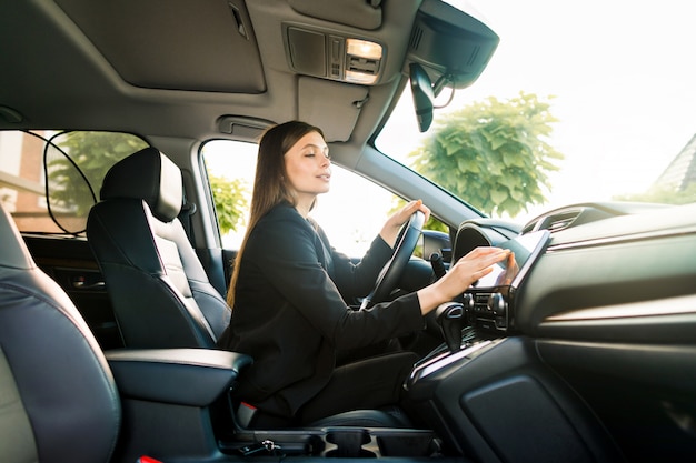 Businesswoman in black suit sits behind the wheel of a premium car and looks the way looking at the monitor of navigation system